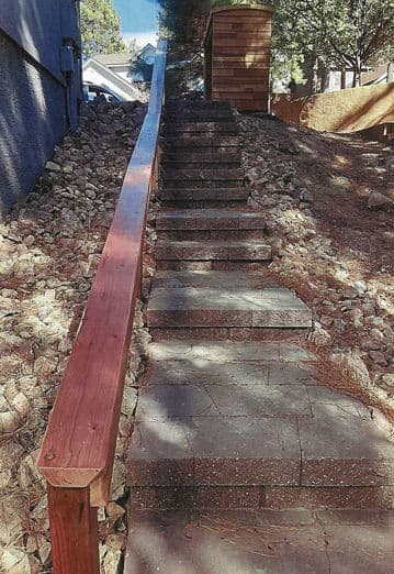 Stone staircase with wooden railing surrounded by rocks and pine needles. Bright daylight setting.