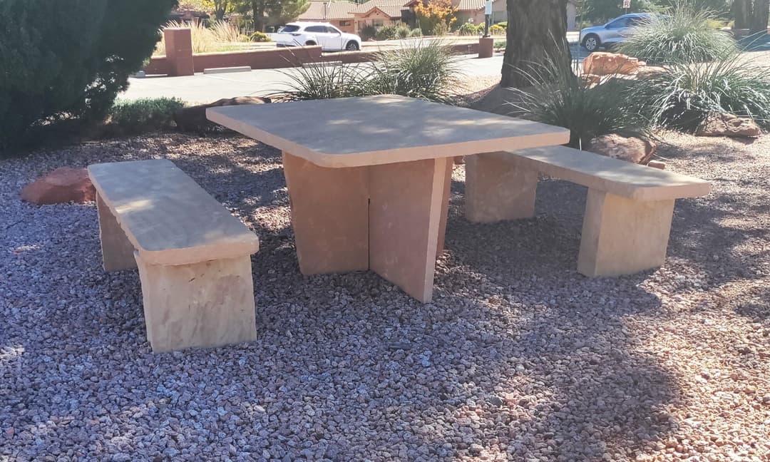 Outdoor stone table and benches in a landscaped area with gravel and greenery.