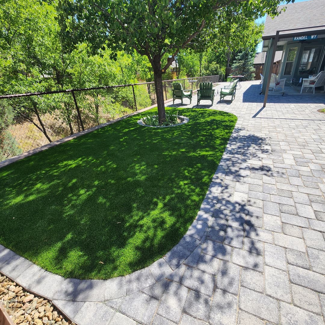 Lush synthetic grass area under a tree with patio furniture and a fenced backyard.