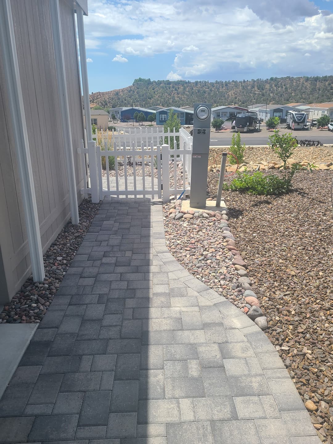 Pathway with pavers leading to a white fence and utility meter in a landscaped area.