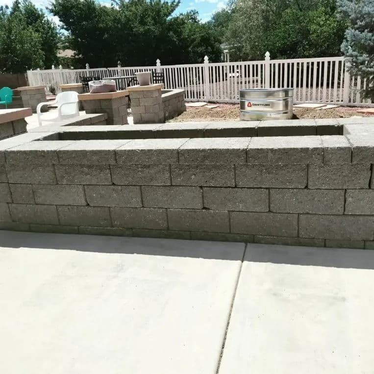 Concrete block wall with fire pit area in a backyard setting, surrounded by greenery.