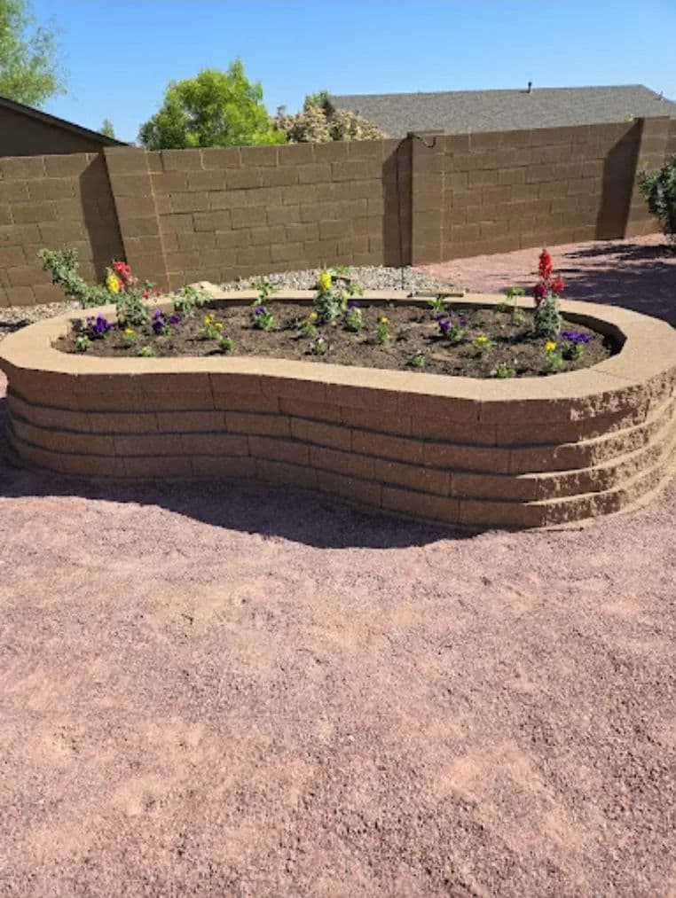 Raised flower bed with colorful plants in a landscaped yard surrounded by a stone wall.