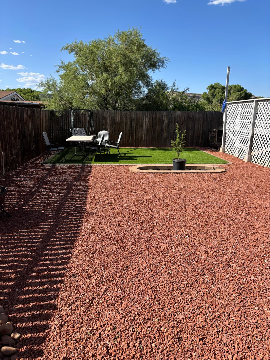 Backyard landscape with red gravel, seating area, and green lawn under a clear blue sky.